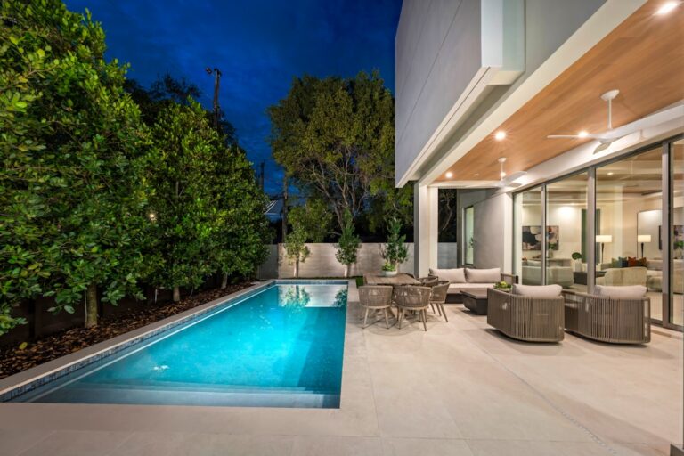 A backyard pool surrounded by a stone patio, lounge chairs, and lush landscaping with a shaded seating area.