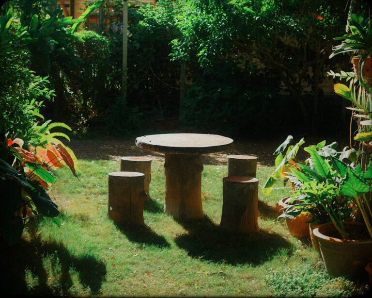 A family relaxing and dining together on a shaded backyard patio with outdoor furniture and lush greenery nearby.