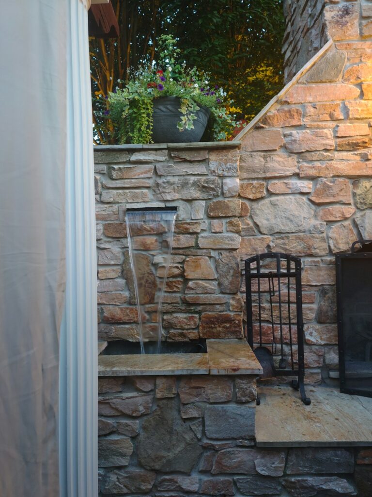 A shaded backyard patio with a bubbling stone fountain, surrounding plants, and a small seating area.
